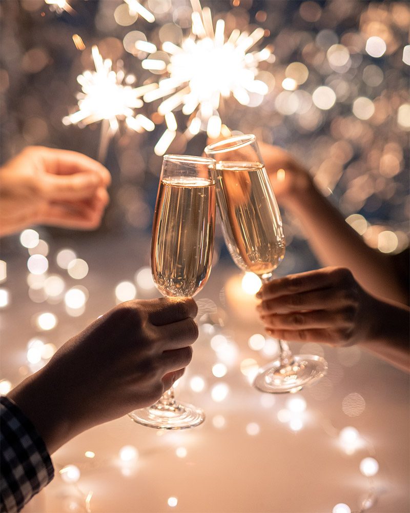 Hands of young couple clinking with flutes of champagne on background of two humans holding sparkling bengal lights